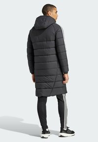 Abrigo largo negro tipo plumas con capucha, que presenta secciones acolchadas horizontales y rayas laterales blancas en unos pantalones negros a juego y zapatillas negras.