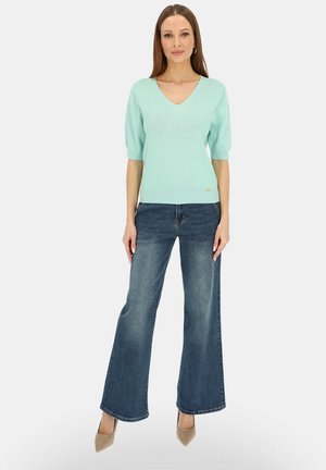 Kobieta stojąca w jasno niebieskim swetrze z dekoltem w serek, szerokich niebieskich dżinsach, beżowych szpilkach i kolczykach kołach na jednolitym tle.