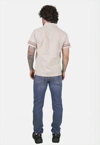 Camicia beige a maniche corte con accenti a contrasto nel polsino, abbinata a jeans blu e sneakers nere. Vista da dietro.