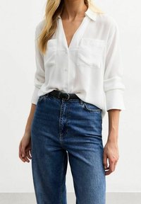 Camicia bianca a maniche lunghe con bottoni e due tasche sul petto, abbinata a jeans in denim blu a vita alta. Tessuto morbido, vestibilità rilassata.
