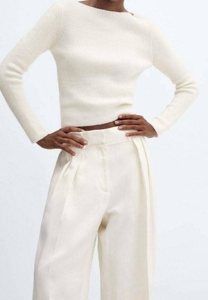 Personne portant un haut ajusté crème côtelé à manches longues avec un pantalon crème taille haute à jambes larges, les mains posées sur les hanches.