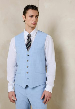 Junger Mann, der eine hellblaue Weste und Hose mit einem weißen Hemd und gestreifter Krawatte trägt, vor einem neutralen Hintergrund steht.