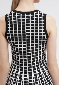 Robe noire sans manches avec motif à carreaux blancs, taille cintrée, vue de dos sur une personne à la peau claire et aux longs cheveux attachés.