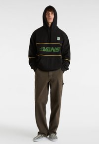 Svart hoodie med grön "VANS"-logotyp, gula ränder och framficka. Kombinerad med bruna byxor och svart-vita rutiga skor.