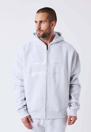 Homme aux cheveux courts et à la barbe, portant un sweat à capuche zippé gris clair et un pantalon de survêtement assorti, regardant sur le côté sur un fond uni.