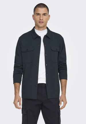 Dark green shirt jacket, two chest pockets, button front, long sleeves, made of textured fabric, worn over a white T-shirt.
