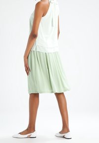 Světle zelená bezrukávová blůza s detailem zavazování na zádech, spárovaná s odpovídající skládací sukní. Modelka má na sobě bílé ploché boty.
