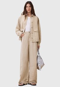 Chaqueta oversized beige y pantalones de pierna ancha de tejido ligero, combinados con una blusa blanca. Sosteniendo un bolso plateado con detalles estructurados.