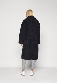 Cappotto lungo nero e peloso con un ampio colletto, vestibilità ampia e tessuto texturizzato; abbinato a leggings grigi e sneakers bianche.