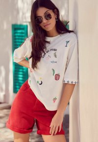 Witte blouse met kleurrijke geborduurde patronen, waaronder fruit en dranken, gecombineerd met rode opgerolde korte broeken. Stijlvolle zonnebril maakt de look compleet.
