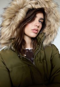 Oliwkowa pikowana kurtka z kapturem z sztucznego futra. Posiada zamek błyskawiczny i brązowe guziki jako akcenty na warstwowej koszuli pod spodem.