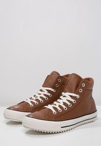 Brown high-top sneakers made of leather with white laces, rubber toe caps, and textured soles. Features silver eyelets and contrasting black stripes.