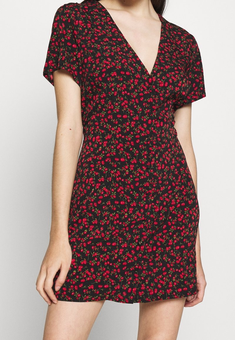 Robe noire à manches courtes avec imprimé cerises rouges, encolure en V et taille cintrée sur une personne aux bras et jambes dénudés.