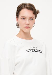 Weißer Sweatshirt mit Perlendetails an den Schultern und einem schwarzen Aufdruck, der "wild heart AWESOME" sagt. Weicher Stoff, lockere Passform.