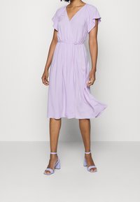 Levandulové midi šaty s krátkými zavinovacími rukávy, výstřihem do V a detailem vázání v pase. Nosené s odpovídajícími levandulovými sandály na blokovém podpatku.