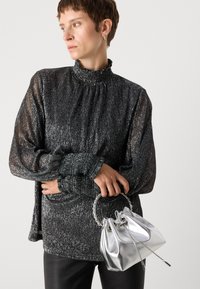 Borsa a mano argentata metallizzata con manico intrecciato, abbinata a una blusa nera a maniche lunghe con texture, caratterizzata da un collo e dei polsini con ruches.