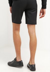 Schwarze sportliche Shorts mit glatter Textur, mittlerer Oberschenkellänge und enger Passform. Getragen mit schwarzen Sneakers. Sichtbare Beine und Hintergrund.