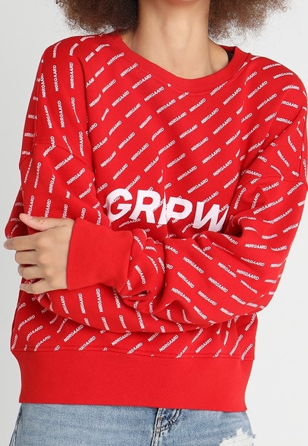 Personne portant un sweat-shirt rouge avec le texte blanc répété "NORGAAARD" et de grandes lettres blanches "GRIPW" partiellement visibles sur la poitrine.