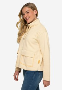 Chaqueta de felpa color crema con un cuello alto, dos bolsillos frontales y acentos de costura naranja. Textura suave y corte relajado.