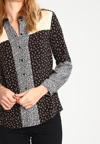 Svart skjorta med knappar och blommönster, med varierande tryck på ärmarna och bröstet. Materialet är lätt och har en slät yta.