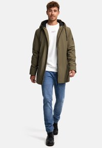 Veste à capuche verte olive avec fermeture éclair, doublure intérieure noire et poches latérales, portée avec un jean bleu clair et des bottes noires.