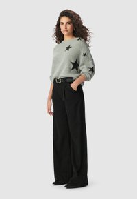 Kobieta z kręconymi włosami, ubrana w szary sweter z czarnymi łatami w kształcie gwiazdy, czarne szerokie spodnie i czarny pasek, stoi z jedną ręką w kieszeni.