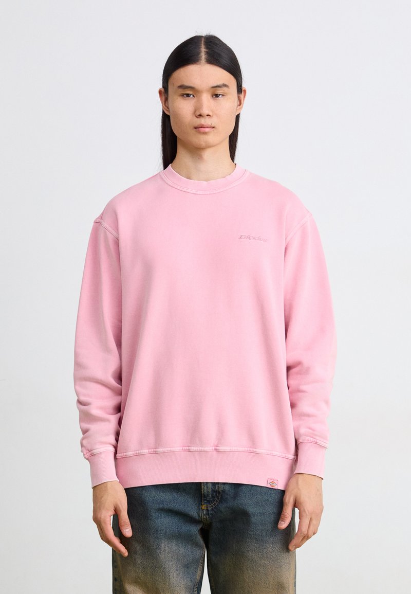 Ροζ φόρμα με στρογγυλή λαιμόκοψη, ριμπ μανσέτες και στρίφωμα. Διαθέτει ένα διακριτικό λογότυπο στο στήθος. Συνδυάζεται με ξεθωριασμένα μπλε τζιν.