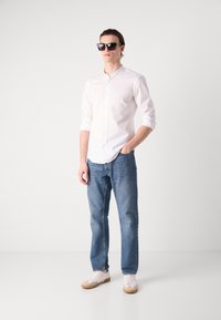 White long-sleeve button-up shirt, blue jeans, and white sneakers. Model wears black square sunglasses. Simple and modern design.