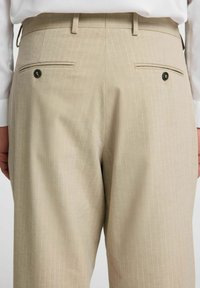 Beżowe spodnie w paski z tylnymi kieszeniami z czarnymi guzikami, dopasowany krój i gładka tekstura materiału.