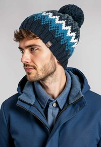 Gebreide winterbeanie in marineblauw, teal en wit met een zigzagpatroon en een pompon bovenop; bevat een klein merketiket aan de zijkant.