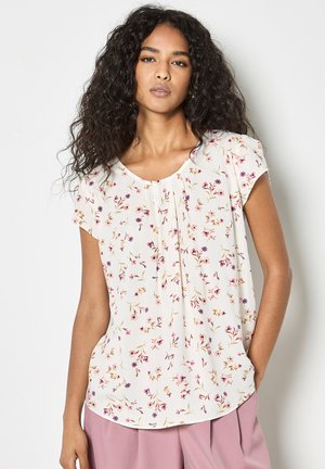 Kobieta z długimi kręconymi włosami, ubrana w białą, kwiecistą bluzkę z krótkim rękawem i różowe plisowane spodnie, stojąca z jedną ręką w kieszeni.