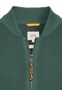 Dunkelgrüner Reißverschluss-Pullover mit geripptem Kragen und strukturiertem Stoff. Verfügt über einen gelben Akzent hinter dem Etikett und einen dekorativen Reißverschlussanhänger.