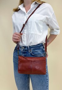 Brun læder crossover taske med en aftagelig rem, der har en glat overflade og en lynlåslukning. Båret med en klassisk hvid skjorte og jeans.