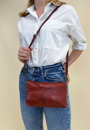 Brown leather crossbody bag with a detachable strap, featuring a smooth texture and a zippered closure. Worn with a classic white shirt and jeans.