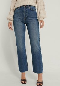 Mujer vestida con jeans azules de pierna recta de cintura alta, un suéter crema de punto trenzado con mangas abullonadas y sandalias negras de tacón abierto.