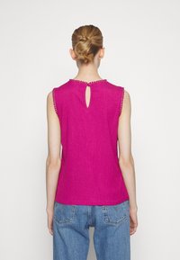 Femme avec un chignon blond portant un haut sans manches magenta avec garniture perlée et dos ajouré, associé à un jean en denim bleu, vue de dos.
