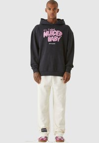Felpa nera con scritta rosa "Si chiama OMICIDIO BABY" sul davanti, abbinata a pantaloni della tuta bianchi e sneakers con dettagli rosa.