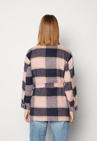 Osoba z prostymi, sięgającymi ramion włosami, nosząca różową i granatową dużą kratkę z paskiem oraz jasnoniebieskie dżinsy, odwrócona tyłem.