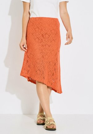Mujer con top blanco, falda asimétrica de encaje naranja y sandalias beige, de pie sobre un fondo blanco.