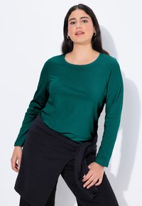 Długi rękaw top w ciemnozielonym materiale z okrągłym dekoltem, w połączeniu z czarną spódnicą typu wrap, z wiązanymi akcentami w talii.