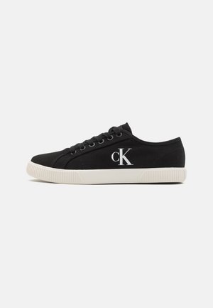 Black canvas sneaker with white rubber sole, lace-up design, and embroidered 'CK' logo on the outer side. Flat profile and textured sole.