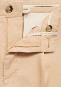 Pantalons beiges clairs avec fermeture éclair et bouton pression. Comprend un tissu texturé en mélange de coton et des accents blancs contrastants.