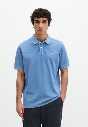 Man met donker krullend haar, gekleed in een lichtblauw poloshirt met korte mouwen en een donkere broek, staande tegen een effen witte achtergrond.