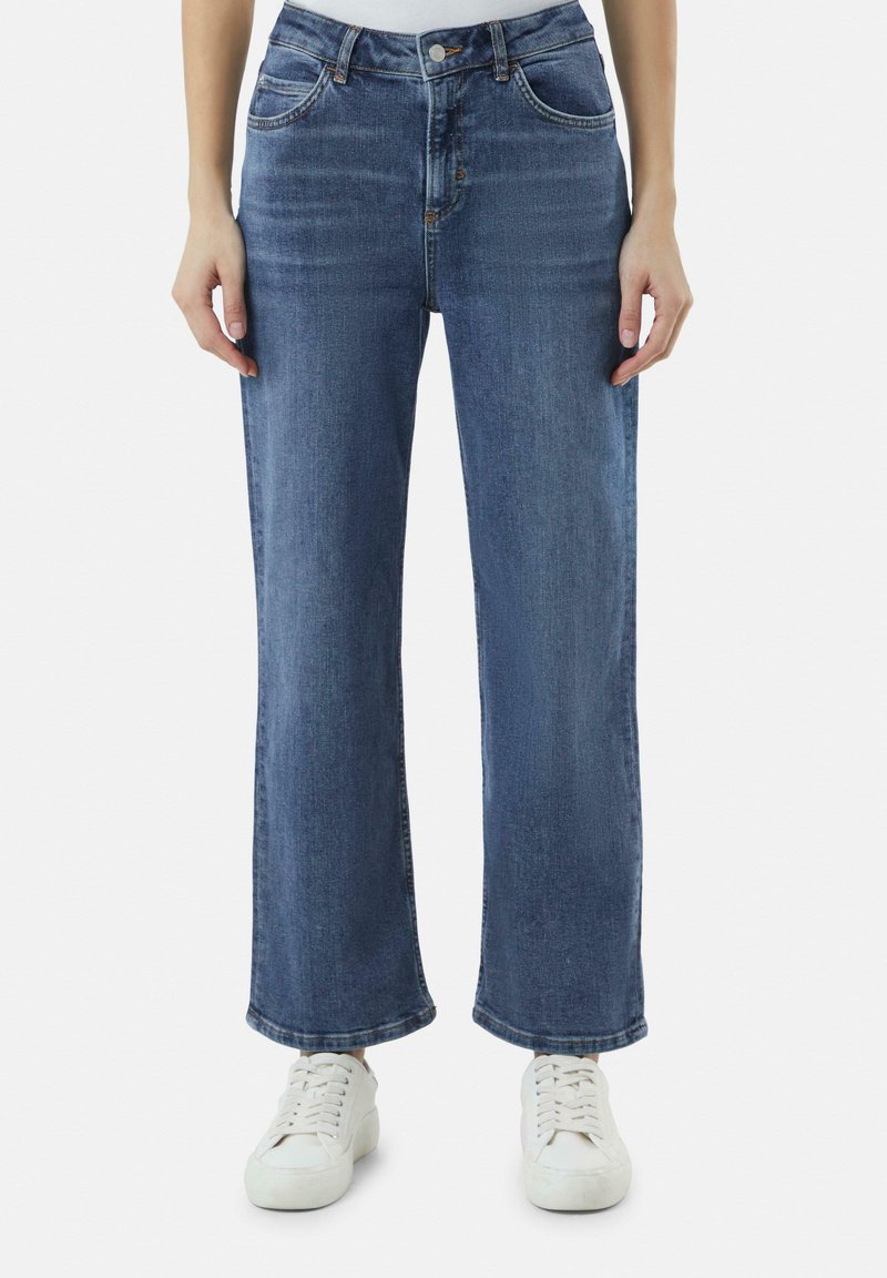 Wide-leg Jeans aus mittlerblauem Denim mit glatter Textur, ausgestattet mit Fronttaschen, einem Knopfverschluss und gesäumten Abschlüssen, kombiniert mit weißen Sneakers.