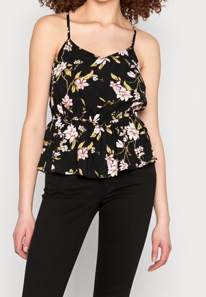 Frau trägt ein schwarzes, ärmelloses Oberteil mit Blumenmuster, dünnen Trägern und einem Volant am Saum, kombiniert mit schwarzen Hosen vor einem schlichten Hintergrund.