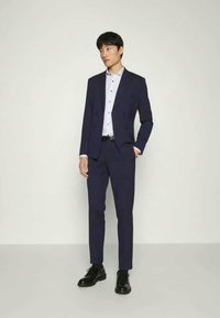 Marineblauer Anzug mit einer doppelt geknöpften Jacke, taillierten Hosen und einem hellen Hemd. Die Schuhe sind aus schwarzem Leder. Glatte Textur, moderner Schnitt.