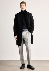 Casaco longo preto de lã com fecho de botões, usado sobre uma camisola de gola alta preta, combinado com calças cinza claras feitas sob medida e sapatos de pele pretos.