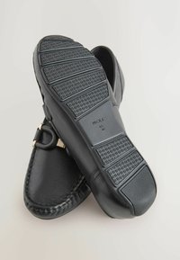Zwarte moccasin-stijl schoenen met een gestructureerde rubberen zool, met stikdetails en een decoratieve gesp. Geplaatst op een lichte achtergrond.