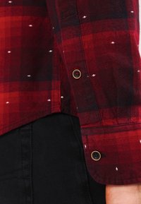 Camisa a cuadros roja y oscura con detalles blancos, con puños abotonados y tela negra de contraste debajo. Material de algodón texturizado.