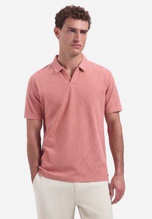 Man met krullend haar, gekleed in een zalmroze korte mouw polo en lichtbeige broek, staand met handen in de zakken.